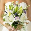 Effortlessly elegant, this Brent Cross wedding bouquet unfolds in the bride's hands like a living expression of hope and artistry. Silk-white Oriental lilies, opening wide, reveal petals dusted with gentle freckles and cradling lustrous golden stamens. Nestled amongst them, lisianthus blossoms bring ruffled textures and soft lavender edges, creating subtle movement and a sense of whimsy that feels right at home in the tranquil corners of Brent Cross's green spaces. Spires of deep purple Veronica burst upward, adding dramatic punctuation beside billowy sprays of baby's breath, which float like a soft mist across the bouquet. Surrounding every bloom is a lush array of verdant foliage-from the broad, shiny leaves to curling lengths of asparagus fern-that cascades gracefully, infusing the bouquet with an air of natural abundance. The bride's thoughtfully manicured hands, finished in a light blush, grasp the gathered stems just above the gentle bloom of her simple strapless dress, itself blurred into the background like a hush before celebration. Each detail evokes not just the joy of a North West London wedding but the personal care, freshness, and warm flair that only florists in Brent Cross can provide, crafting arrangements that remain timeless in photographs and memory alike.
