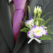This boutonnière, prepared by Brent Cross Flowers with the calm confidence of an experienced hand, sits just above the heart on a dark suit and speaks of considered elegance. The central bloom is a white lisianthus edged in vibrant purple, its silky, slightly ruffled petals forming a soft, luminous cup that draws the eye; three upright lisianthus buds behind it suggest new beginnings, while a single stem of deep indigo veronica adds a refined vertical line. Around them, slim blades of fresh, dark green foliage cradle the flowers and provide a cool, earthy sheen; wisps of ornamental grass lend a touch of movement, and tiny sprays of gypsophila scatter like gentle confetti, brightening the composition without overpowering it. The bouquet is tied with a crisp white ribbon whose neat knot mirrors the arrangement's restrained formality, and the faint floral perfume-green, clean, and reassuring-settles pleasantly for hours. Natural daylight glances across the petal edges, the purple rim sharpening the silhouette and giving subtle theatricality against the wearer's magenta tie. As a local piece suited for Brent Cross and neighbouring NW London areas-from the shopping centre to Claremont Park and beyond-this boutonnière is designed to withstand a long celebratory day: careful wiring and selection of fresh stems ensure it keeps its posture and presence. It's a detail chosen for weddings, anniversaries, or any event where a quiet, personal touch matters: an emblem of taste that complements the suit and honors the moment with understated beauty.