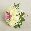 A delicately styled petite bouquet is captured from a slightly elevated angle, resting on a neutral light beige fabric that looks like finely woven linen, its subtle texture adding a calm, natural backdrop. At the centre, five creamy white roses open in soft, rounded layers, their ivory petals forming generous, spiralling cups that suggest classic romance and careful, early-morning conditioning by an experienced florist. Nestled toward the front-left is a single fuchsia-pink rose, bold yet graceful, providing a confident splash of colour that draws the eye and gives the arrangement personality-ideal for a standout corsage at a Brent Cross school prom or an elegant boutonnière for a wedding at a nearby hotel. Airy sprigs of white baby's breath lace through the design, their tiny blossoms creating a mist-like veil that lightens the composition and adds an almost ethereal, bridal quality. Around the blooms, deep green, pointed foliage-likely ruscus or salal-frames the flowers, the leaves smooth and slightly glossy, adding depth, contrast, and a sense of freshness you might associate with a walk through Brent Cross's tree-lined residential streets. The stems are neatly bound into a compact cluster and wrapped in a bright pink satin ribbon, the surface shining gently under soft, even lighting. Over this ribbon, fine rows of small white pearls are carefully arranged in horizontal bands, their subtle iridescence suggesting thoughtful craftsmanship and a touch of vintage glamour. The overall scene feels intimate, romantic, and composed for a special occasion, ideal for same-day gift delivery around Brent Cross, Golders Green, or Cricklewood when a refined, small-scale floral piece is needed to mark a meaningful moment.
