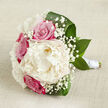 On a backdrop of pale, textured beige fabric, reminiscent of fine linen laid out on a bridal preparation table, a hand-tied bouquet rests in a horizontal pose, filled with soft romance and quiet luxury. At the very centre sits a generous creamy white peony, fully open and lush, its countless ruffled petals creating a deep, cushion-like bloom that radiates gentle light. Around it, several fuchsia-pink roses gather in close, their tightly spiralled petals and rich colour adding a classic romantic intensity, as if echoing the excitement of couples getting ready for a ceremony close to Brent Cross. Light sprays of white gypsophila thread between the larger blooms, the tiny, cloudlike flowers giving the arrangement a floating, almost magical texture that softens every edge. Framing this floral heart are broad, dark green leaves, slightly curled and polished, wrapping around the bouquet in smooth arcs that lend structure and freshness, like a patch of green tucked between the busy roads and retail centres nearby. The stems are neatly trimmed and bound with a pristine white satin ribbon, carefully wound to create a sleek handle that hints at comfortable, all-day carrying-ideal for a bride walking from a local home to a nearby venue. Soft, even lighting bathes the entire design, revealing subtle colour transitions from deep fuchsia through gentle blush to pure cream, and capturing the natural sheen of each petal and leaf. The bouquet communicates calm happiness and refined celebration, crafted by a florist who understands Brent Cross couples seeking something timeless yet modern for their wedding or special occasion.