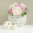 A romantic bouquet of ivory peonies and vibrant fuchsia-pink roses is captured in serene, diffused light, resting on a pristine white tabletop that reflects the arrangement's soft shadows. At the heart of the display, the peonies unfold in luxurious layers, their creamy petals curling and overlapping like fine fabric, suggesting both delicacy and quiet opulence. Around them, the roses bring vivid energy, displaying rich pink tones that deepen toward the center, their velvety appearance hinting at a subtle, classic rose fragrance. Scattered between the larger flowers are airy sprigs of white baby's breath, each stem carrying clusters of tiny blooms that create the impression of a light, floral haze surrounding the main blossoms. The bouquet is framed by smooth, broad green leaves that curve gently outward from the base, creating a dome-like, almost bridal profile ideal for weddings and celebrations across Brent Cross, from intimate gatherings near Hendon Way to receptions close to the shopping centre. The flowers are gathered into a distinctive white vase with an open, lattice-style structure, as if made from intertwined, painted branches or fine rattan. This see-through, basket-like design allows glimpses of the stems within and adds a sculptural, modern touch that contrasts beautifully with the softness of the petals. Positioned in front of the vase are two pale, woven decorative orbs, one slightly larger, both echoing the vase's texture and reinforcing the theme of organic craftsmanship. The background is a warm, neutral beige with subtle texture, similar to finely woven fabric, providing a calm backdrop that makes the colours of the flowers gently stand out. The entire arrangement feels poised and celebratory, perfectly suited for bridal table decor or same-day congratulations delivered by a local Brent Cross florist.