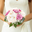There is a tranquil elegance to this bouquet, the kind that might accompany a bride slipping quietly into a ceremony at a Brent Cross hotel or synagogue, everything around her suddenly hushed. The arrangement is intentionally compact, drawn close into a hand-tied cluster that rests easily in her grasp. Dominating the centre, grand creamy peonies unfold with serene confidence, their petals unfurling in soft, rounded layers like whispers of silk. Their off-white hue leans towards warm cream, creating a gentle glow that feels comforting and timeless. Around them, roses in muted dusty pink and cool soft violet weave through the design, each bloom at a perfect stage of openness, with visible spirals of petals that gently loosen towards the edges. These touches of colour introduce subtle vibrancy, shaping a delicate interplay between romance and restraint. Between the larger flowers, airy wisps of gypsophila peep out, their tiny white florets bringing an almost weightless texture that recalls a scattering of fine bridal lace, similar to glimpses of intricate fabric seen in wedding boutiques around Brent Cross. Polished green leaves cradle the bouquet from beneath and around the sides, their deep hue enhancing the pastels while offering a natural frame. The foliage's smooth surfaces catch small glints of light, giving the design movement and depth. Everything about the bouquet feels intimate and personalised, as though crafted with a clear understanding of the couple's style and story. It exudes calm, harmony, and quiet joy, perfectly suited to weddings across Brent Cross, Golders Green, and Cricklewood, where fresh, carefully chosen blooms help mark the start of a shared life.