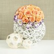 A graceful floral centerpiece is displayed as though ready to be carried into a wedding venue or set on a celebration table in Brent Cross, the arrangement balancing romance, texture, and contemporary design. The eye is first drawn to a perfectly rounded mound of roses in soft peach and apricot tones, the blooms packed closely together to form a lush, velvety dome. Each rose is fully open, revealing layers of gently curled petals that create depth and a subtle play of light and shadow across the rounded surface. Nestled around the base of this rose sphere, a band of white lisianthus blooms encircles the arrangement, their delicately ruffled petals each tipped with a vivid ring of deep violet or purple. This bold edging adds a sophisticated contrast to the gentle peach tones above. Between the lisianthus blossoms, fine sprays of white gypsophila are woven in, their tiny star-like flowers softening the transition and adding an airy, almost cloud-like feeling. Supporting the arrangement is a white woven vessel with a bulbous, spherical silhouette, crafted from interlacing strands of rattan or wicker that form an open lattice. This sculptural vase, with its crisscrossing lines and pockets of shadow, feels handcrafted yet modern, well-suited to stylish homes and event spaces around Brent Cross and nearby Golders Green. Resting on the same smooth white surface in front of the vase are two coordinating decorative spheres, one larger and one smaller, created from slim, pale wooden strips. Their open, woven design mirrors the container and gently leads the gaze back to the flowers. A soft, beige, fabric-textured background completes the image, lending warmth without distraction. The result is a scene of quiet sophistication and heartfelt celebration, ideal for conveying the look of wedding table flowers or elegant home décor in the Brent Cross area.