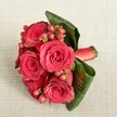 A luxurious cluster of deep red roses fills the centre of the image, gathered into a compact, hand-tied bouquet and resting on a soft, light beige fabric with a subtle woven texture, like fine linen laid over a table in a Brent Cross townhouse. Each rose is fully open yet still structured, with velvety petals that spiral from rich crimson hearts to slightly softer scarlet edges, suggesting the fullness of mid-bloom rather than the fragility of fading flowers. Nestled between the roses are glossy, rounded Hypericum berries in shades of red and reddish-pink, their smooth skins catching the gentle, even light and creating tiny highlights that contrast with the plush matte of the petals. Around the base, broad, dark green leaves are carefully curved and wrapped, forming an elegant collar that cradles the blooms and adds sculptural depth; you can almost feel the cool, slightly waxy surface beneath your fingertips. A rich red satin ribbon holds the stems tightly together, a neat band visible at one side, its sheen echoing the elegance of evening wear at a wedding reception near Hendon or Golders Green. The neutral background, softly lit without harsh shadow, makes the deep reds and lush greens appear even more vivid and romantic, as if ready to be lifted and handed to a partner at an anniversary dinner in a Brent Cross restaurant. The composition, viewed from a slightly elevated angle, feels intimate and intentional, capturing a bouquet perfect for a proposal, a Valentine's Day surprise, or a refined bridal accessory with timeless, classic beauty.