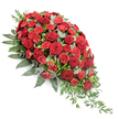 In this image, a traditional yet gracefully modern funeral spray is shown against a bright white background, its elegant diagonal shape and rich colours standing out with clarity. The arrangement forms an elongated teardrop or casket spray, full and rounded at the upper left before tapering into a slender, trailing point at the lower right, suggesting a gentle, downward cascade. The central focus is a collection of deep red roses, their velvety petals fully open and layered, creating soft, cupped blooms that evoke love, respect and heartfelt remembrance. Nestled closely among them are clusters of red spray carnations, their countless ruffled petals providing a delicate, cloud-like texture that fills the spaces between the larger roses. This interplay of blooms creates a dense, luxurious field of crimson shades. Supporting and framing the flowers is an arrangement of lush greenery. Broad, dark green leaves curve around the outer edges, providing structure and a strong silhouette, while rounded eucalyptus leaves in a soft, silvery green are woven through the centre, cooling and balancing the warm reds with their gentle hue. From the narrow tip of the spray, fine strands of lighter trailing foliage extend outward, enhancing the cascading character and adding a natural, almost river-like flow to the piece. The overall atmosphere is one of quiet beauty and dignified sorrow, well-suited for a funeral service or memorial gathering. Imagined in Brent Cross, it might be seen resting on a casket in a chapel near the North Circular or brought by same-day delivery to a family home in nearby Hendon or Temple Fortune, a thoughtful floral tribute that honours memory with grace and care.