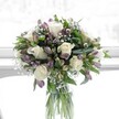 A rounded bouquet of creamy white roses, lavender alstroemeria, and airy baby's breath stands in a clear cylindrical glass vase, softly lit as if placed near a bright window in a Brent Cross home. The fully opened white roses are the stars of the arrangement, their velvety petals spiralling outward in perfect layers, creating a gentle, romantic focal point. Around them, clusters of alstroemeria in muted shades of lilac and dusky purple offer a graceful contrast, their slender buds and partially opened blooms streaked with fine, darker veining that adds depth and intrigue. Between the larger flowers, tiny cloud-like sprigs of white gypsophila weave in and out, giving the bouquet an almost misty, weightless quality that lightens the entire design. Rich green foliage, both broad and glossy and more slender and linear, frames the flowers and strengthens the rounded silhouette, the leaves catching hints of light to show their fresh texture. Through the transparent glass, the long, neatly trimmed stems are clearly visible, submerged in clean water that suggests same-day preparation and delivery, much like arrangements travelling out across Brent Cross, Hendon, and Golders Green. The background is softly diffused and pale, free of clutter, so that the eye rests fully on the interplay of white, green, and purple. The overall mood is one of calm, refined elegance and quiet optimism, evoking a sense of fresh beginnings after gentle spring rain. It feels like the kind of bouquet chosen to bring comfort after a difficult time, to mark a thoughtful anniversary, or to brighten a hallway near Brent Cross Shopping Centre, its refined palette and careful craftsmanship speaking for someone who wants to express love, sympathy, or simple, heartfelt appreciation without saying a word.