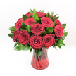 A studio-style photograph shows a classic, luxurious bouquet of deep red roses arranged in a rich ruby glass vase, placed alone against a crisp white background so every detail is clear. Around a dozen velvety crimson blooms form a full, rounded dome, each rose at perfect maturity with spiralling petals and gently furled centres that suggest romance just about to unfold. Between the blossoms, polished dark green leaves, similar to Ruscus foliage, peek through and extend slightly beyond the lower edge of the bouquet, giving the design structure and lush contrast. The vase itself is a translucent red glass, softly fluted with a subtle ribbed or swirling texture that catches the light and echoes the colour of the blooms above. Through the tinted glass, you can make out the dark vertical lines of the rose stems, neatly gathered and crossing in a natural pattern, adding depth and a sense of hand-crafted care. Bright, even lighting washes across the arrangement, intensifying the ruby hues and highlighting each petal's soft texture, as if the flowers have just arrived from a morning delivery. The overall effect is timeless and passionate, ideal for a Brent Cross couple celebrating an anniversary at home near the shopping centre, or for a dramatic Valentine's Day surprise sent to an office overlooking the North Circular. Though the setting is minimal, the bouquet feels abundant and heartfelt, the kind of red rose arrangement a skilled local florist would create when asked for something that speaks clearly of love, devotion, and special-occasion elegance without any unnecessary embellishment.