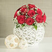 At the heart of the image stands an elegant arrangement of red roses, their petals a rich, saturated crimson that spirals inward with velvety precision. The roses are grouped so closely that their heads form a neat, rounded mound, suggesting a dozen or so carefully selected stems chosen at the same stage of bloom. They are framed by a delicate collar of white baby's breath, each stem branching into clusters of tiny, snow-like blossoms that create a soft, clouded edge to the bouquet. Subtle touches of dark green leaves emerge from beneath the flowers, adding depth without stealing focus from the interplay between red and white. This crowned bouquet is nestled into a contemporary white sphere-shaped vase, its surface made from fine rods woven into a crisscross pattern that recalls hand-crafted wicker yet feels sleek and modern.
The vase rests on a pristine white table, where the open structure allows pockets of light to fall through, sketching faint patterns that echo the lattice design. Two decorative balls sit to one side, one larger, one smaller, made from thinner, cream-toned strands that look like pale rattan or dried vine. Their neutral shade harmonises with the vase and tabletop while providing a gentle contrast in texture, like stylish accents you might spot in a Brent Cross flat overlooking the North Circular or a newly renovated home near Clitterhouse Playing Fields. The background is a soft, neutral wall in beige or light grey, free of clutter so that all attention centres on the flowers and their sculptural surroundings. The atmosphere is calm, romantic, and refined, perfectly suited to a wedding table arrangement, proposal dinner, or heartfelt gift, whether placed in a restaurant near Brent Cross or delivered as a surprise to a loved one at home.