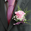 In this photograph, a romantic wedding boutonniere is captured up close, pinned precisely to the lapel of a charcoal-grey suit, the sort of attire you might see at a ceremony near Brent Cross or a smart celebration along the A41. At the centre of the composition is a single blush-pink rose, its petals silky and gently curved, unfolding from a soft, creamy core into a more defined rose-pink edge. The bloom is at that perfect stage of openness-full enough to feel generous, but still structured and tidy, with a sense of quiet poise. Nestled around the base of the rose are fine clusters of white gypsophila, their miniature blossoms forming a delicate, airy cushion that lightens the overall look and adds texture without overwhelming the central flower. A few slender, fresh green leaves and stems extend just beyond the edges of the rose, providing an easy, natural frame that suggests movement and life, like a small echo of the leafy residential streets off Highfield Avenue. The stems of the boutonniere are neatly bound with a slim satin ribbon in a dusky lilac-pink shade, wrapped smoothly and held in place with invisible care, the subtle shine of the ribbon catching the ambient light. The dark suit fabric beneath is clearly defined, offering a tailored contrast to the softness of the petals and baby's breath. Together, these elements create a look that is elegant yet understated-perfect for a groom, groomsman, or prom attendee in Brent Cross who wants a floral accent that whispers romance rather than shouts it. The overall mood is one of tenderness and intention, as though each stem and fold of ribbon has been chosen with a particular promise in mind.