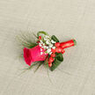 Carefully composed with the certainty of an experienced hand, this boutonniere centers on a saturated magenta rose whose velvety petals are coaxed into a poised, half-open bloom. Tiny sprays of white gypsophila float around the main flower like soft, bright mist, their fragile clusters creating contrast and lending an air of nostalgia. Glossy red hypericum berries nestle discreetly among the tiny white flowers, offering concentrated pops of crimson that mirror the satin ribbon binding the stems. The foliage is layered for movement and form: fine, grass-like sprigs provide feathered texture and an informal breeze-like silhouette, while broader, folded green leaves give the piece an architectural backbone and a refined edge. The stems are wrapped in bright red satin ribbon, tightly and neatly finished so the boutonniere feels secure and sits flat on a lapel. The neutral woven background in the image allows each colour and texture to be read clearly-the rose's deep pink, the pure white of baby's breath, the glossy berries and varied greens all hold their place in the design. You can imagine the soft fragrance of rose with an undernote of fresh foliage, subtle but present. Made with local sensibility for Brent Cross and deliveries into nearby Golders Green and the shopping centre, this is the sort of piece Brent Cross Flowers would prepare for a summer wedding, prom, or anniversary-elegant, celebratory, and designed to be felt as much as seen.