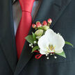 A close, intimate view of a classic boutonnière pinned to the left lapel of a charcoal-black suit, where floristry and menswear meet in tasteful harmony. The focal point is a single white Phalaenopsis orchid, its broad, velvety petals luminous under soft light and centered by a yellow-gold patterned lip that reads like a small sunlit crest. Surrounding the bloom, deep green glossy leaves form a protective frame, their texture smooth and slightly reflective; delicate sprays of gypsophila drift outward like a pale confetti, introducing a whisper-soft texture and an air of celebration. Rounded hypericum berries - blushing coral to warm red - cluster near the base, their tactile presence and colour quietly echoing the solid red satin tie knotted sharply at the throat. A slender loop of matching red ribbon secures the stems and ties the ensemble to the tie's confident hue. Visible beneath, a subtle light blue or grey checked dress shirt provides a cool, patterned backdrop that highlights the boutonnière's clarity. This arrangement speaks of considered craftsmanship and fresh materials - the kind of finishing touch men ask for when attending a Brent Cross wedding, a reception at the shopping centre, or a marquee event near the North Circular. Prepared by local hands and offered by Brent Cross Flowers for neighbours across Cricklewood and Golders Green, the boutonnière balances modern simplicity with timeless formality.
