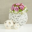 On a clean white tabletop, a single, thoughtfully designed centerpiece suggests the calm before a celebration in one of Brent Cross's wedding venues. The arrangement is low and rounded, composed of a lush gathering of roses in blush pink and soft lavender tones. Each rose is open and expressive, with petals that spiral outward and deepen slightly in colour at the edges, creating gentle shadows and a feeling of depth. Between the roses, bright green button chrysanthemums are set like small, neatly clipped garden mounds, bringing a vivid splash of green that keeps the palette fresh and modern. Around the lower edge of the bouquet, fine white filler flowers appear in tiny sprays, giving the composition a delicate, almost airy fringe that blurs the transition into the container. The flowers are held within a spherical white vase fashioned from slim, rattan-style twigs, woven into an open, basket-like structure. This contemporary container has a lightness that matches the easy elegance often seen in Brent Cross homes and event spaces, combining natural inspiration with clean design. To the left of the vase, two decorative wicker spheres made from pale, interlaced strands rest casually on the white surface, one larger, one smaller, their shapes echoing the vase's woven pattern and adding subtle visual interest. The background is softly blurred in a warm beige tone, likely a textured linen or burlap fabric, which provides a neutral, inviting backdrop and allows the pinks, greens, and whites to stand out clearly. Soft, even lighting falls across the scene, highlighting the velvety rose petals and the intricate weave of the wicker, making this arrangement feel like an ideal wedding table decoration for receptions and special occasions across Brent Cross, thoughtfully crafted by Brent Cross Flowers.