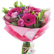 This bouquet reads like a bright, uplifting moment captured in floristry-fuchsia gerbera daisies with almost-black centers stand proud at the heart, their velvety, densely packed petals radiating energy and cheer. Around them, soft pink roses gently unfurl in varied stages, from tightly coiled buds to fuller blooms with layered, satiny petals, giving the composition a whispered romance beneath its bold headline. Sprays of deep purple-blue filler flowers-statice or limonium-are tucked between stems, adding fine-grained texture and a subtle cool counterpoint that draws the eye inward. Lush green foliage with broad, smooth leaves forms a supportive backdrop, the glossy and matte greens grounding the intense pinks and lifting the bouquet's natural silhouette. All of this is swaddled in crinkled, semi-sheer pink wrapping that graduates from a pale, almost translucent halo at the top to a richer rose-pink toward the base; the wrap catches studio light in soft highlights and creates a voluminous collar for the flowers. The stems sit visibly in a clear cylindrical glass vase, water glinting where a rustic twine is tied around the neck; a small white rectangular tag hangs from the twine, printed with a simple pink-and-green rose emblem that signals a florist's touch. The scent is hinted at-gentle, sweet, freshly cut-like a morning at a neighbourhood flower stall. As a local florist might suggest, this arrangement suits birthdays, thank-you gestures, or to brighten a new home in Brent Cross, Cricklewood or nearby Golders Green, where its joyful, confident palette will feel instantly familiar and warmly received.