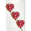 In this serene funeral tribute, three heart-shaped arrangements of roses are displayed in a cascading diagonal, their shapes clearly defined against a bright, uncluttered white wall. Each heart is a tapestry of roses in deep crimson red, vivid fuchsia, and soft pastel pink, the flowers closely packed so that petals gently press against one another, creating a plush, luxurious texture. The varying stages of bloom-from small, tight buds to fully expanded roses with layered, spiralling centres-add depth and a quiet sense of movement, as if the hearts themselves were breathing with memory. Slim vines of fresh ivy trace the outline of each heart, the small, sharply pointed green leaves softening the edges, while longer strands fall between the hearts, binding all three together in a continuous chain. The palette of reds and pinks conveys both sorrow and tenderness, making the design particularly suitable as a tribute for a beloved partner, devoted parent, or close friend. The clean white backdrop, similar to the calm interiors of chapels and service rooms found near Brent Cross and along Hendon Way, ensures the flowers remain the central focus. Gentle, even lighting removes harsh contrasts, instead casting a soft shadow to the right that enhances the three-dimensional quality of the piece. Crafted with the practiced skill of Brent Cross Flowers, this triple-heart arrangement feels like a final love letter written in petals, ideal for funerals or memorials where families wish to express enduring affection, gratitude, and the belief that the bonds of the heart remain unbroken even after goodbye.