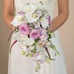 A detailed, intimate shot highlights the artistry of a Brent Cross bouquet as a bride holds a lavish, cascading arrangement brimming with romance and style. Dominating the composition, broad white orchids glisten beside lush pink and lavender roses-some tightly budded, others adorned with full, velvety blooms. Sprays of gypsophila weave through the florals, adding a cloud-like softness, while slender magenta amaranthus and trailing greenery lend movement and contemporary drama. Glimpses of the bride's lace-trimmed ivory gown and her gentle touch evoke the quiet excitement found on wedding mornings near Claremont Road. The background, serene and understated, draws the eye forward to the vibrant intricacies of each bloom. Perfect for spring or summer ceremonies across Brent Cross, this bouquet reflects the skill and passion of local florists and the joy of beginning a new journey in North West London.