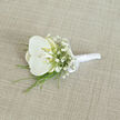 A refined orchid boutonnière rests at a gentle angle on a pale, linen-like fabric, as if waiting to be pinned onto a freshly pressed suit for a Brent Cross wedding. At its heart is a single white orchid bloom, generous and perfectly formed, with broad petals that appear smooth and cool to the touch, like polished porcelain. The center of the orchid reveals a soft blush of creamy yellow deep in the throat, a subtle glow that brings warmth and dimension to the otherwise pure white flower. Encircling this focal bloom are clusters of airy white baby's breath, each stem branching into dozens of tiny blossoms that together form a weightless, cloud-like halo. Fine strands of vivid green foliage slip out from underneath the floral cluster-slender blades that might be decorative grasses or delicate fern fronds-adding a fresh, lively contrast and suggesting a whisper of woodland greenery, not unlike the calm found in nearby Brent Reservoir. The stems are neatly aligned and expertly wrapped in smooth white satin ribbon, creating a polished handle with a faint, elegant sheen. Near the top of this wrapping, a small, perfectly tied satin bow offers a final touch of classical detail, hinting at careful, experienced craftsmanship. The background fabric, a light beige with a visible weave, introduces gentle texture without competing for attention. Soft, even lighting reveals the richness of each surface-the orchid's velvety petals, the tiny buds of gypsophila, the crisp ribbon edges-conveying an atmosphere of serene sophistication. It feels like a timeless boutonnière for a groom or honoured guest, suited to intimate ceremonies in and around Brent Cross.