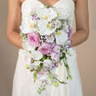 In this tender bridal portrait, the camera focuses on the bride's midsection, where a smooth white strapless gown forms a calm backdrop for an opulent, cascading bouquet. The bouquet is an elegant teardrop that begins just below her waist and flows downward, almost like a waterfall of petals. Large white Phalaenopsis orchids occupy pride of place, their wide, curved petals silky and unblemished, surrounding a soft yellow-orange centre that brings a hint of golden light into the composition. Around these orchids, clusters of vivid pink roses and softer, muted lavender-pink garden roses bloom profusely, building gentle mounds of texture and colour that speak of romance and devotion. Smaller white blossoms, likely lisianthus with faint purple edges, nestle in between, echoing the lavender tones and helping to blend the different shades. Feathery tufts of white baby's breath are scattered throughout, adding airiness and a sense of delicacy, as if the bouquet has been lightly dusted with mist. Here and there, slender stems of light lavender stock emerge, offering both subtle height and the impression of a cool, fresh scent one might notice while stepping out of Brent Cross Underground Station on a mild day. The entire arrangement narrows as it descends, creating a refined teardrop that feels both structured and naturally flowing, ideal for gliding down the aisle in a North West London wedding venue. This photograph would be perfect for illustrating bridal bouquet trends, orchid and rose combinations, and romantic wedding flowers from a local florist serving Brent Cross, Golders Green, and the neighbouring communities.