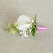 A delicate orchid boutonniere is laid horizontally on a softly textured, oat-coloured linen background, the neutral fabric providing a calm, understated stage for the flowers. At the centre sits a single white Phalaenopsis orchid, its broad petals smooth and luminous, curving gently outward like folded silk. The heart of the bloom reveals intricate details of soft yellow and a whisper of pale pink, the throat of the flower drawing the eye inward with its subtle patterning. Around the base of the orchid, tiny white baby's breath blossoms cluster in cloud-like sprays, their fine stems and minute blooms creating an airy, romantic halo. A few slim, vibrant green leaves peek from behind, offering a fresh contrast to the white petals and creamy backdrop. To one side, a short lateral stem holds several closed, lime-green orchid buds, their rounded forms suggesting the quiet promise of future blossoms. The stems are gathered and wrapped in a bright fuchsia satin ribbon, tied into a neat, elegant bow whose glossy sheen catches the light and adds a vivid pop of celebratory colour. The lighting is soft and even, revealing the velvety texture of the orchid, the delicate granularity of the baby's breath, and the gentle weave of the linen beneath. The overall mood is one of refined simplicity and intimate elegance, perfectly suited to a groom's lapel, a prom date's jacket, or a special guest's outfit at a Brent Cross celebration, whether it's a registry office wedding, a local hotel reception, or an evening event overlooking nearby parks and tree-lined streets.