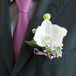 This image captures a sophisticated orchid boutonniere resting against the dark lapel of a well-cut suit, the sort you might see at a stylish wedding reception just a short drive from Brent Cross. A single white orchid takes centre stage, its petals broad and slightly cupped, glowing softly with a hint of ivory warmth. Their smooth surface suggests the cool touch of fine porcelain, and the flower's centre is delicately detailed, drawing the gaze inward. Surrounding the orchid, silvery-green foliage fans out with elegant restraint, the leaves narrow and polished, framing the bloom without overwhelming it. Intertwined among the greenery are slender lilac sprigs and tiny lavender flowers, their pastel tones bringing a gentle whisper of colour, like a soft echo of the nearby tie. Small white gypsophila-style buds are sprinkled throughout, adding a light, cloud-like texture that keeps the design fresh and modern. The stems are bound with a narrow violet ribbon, gleaming subtly as it wraps tightly around the base, mirroring the mauve and lilac shades of the tie beneath the lapel. The charcoal suit jacket provides a strong, masculine contrast, making the orchid seem almost luminous in comparison. Soft, natural light falls across the scene, picking out the veins in each petal and the fine weave of the fabric, without harsh shadows. The composition feels intentional and balanced, perfectly suited for a day of vows, photographs, and toasts-whether at a local Brent Cross venue, a synagogue nearby, or a family gathering just off the North Circular. It's a small but expressive detail, capturing both celebration and quiet sentiment.