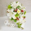 A graceful floral centerpiece rests on a smooth white tabletop, its refined style perfectly suited to a modern wedding reception or engagement dinner in Brent Cross. The flowers emerge from a round white woven basket, the wicker forming a soft lattice that cradles the composition and adds gentle texture. At the core, large white lilies open in full trumpet shapes, their petals silky and luminous, touched with faint speckling that draws attention to the central yellow anthers dusted with pollen. Slightly above them, a single cream-yellow rose, still somewhat budded, stands as a subtle focal point, its layered petals promising to unfurl further. Drifting from the right-hand side, a cascade of white phalaenopsis orchids trails elegantly downward, each bloom perfectly poised, creating a flowing, waterfall-like line that suggests movement and elongates the design. Around these feature blooms, an abundance of green foliage and textural accents fills the basket: feathery sprigs, slender grass-like leaves, and bright lime-green pom-pom flowers that add a plush, contemporary touch. Threaded delicately between the larger flowers, tiny white blossoms reminiscent of baby's breath create a fine, cloud-like veil, while clusters of rich red berries offer vibrant pops of colour, similar to festive arrangements delivered around Brent Cross at Christmas and special occasions. To the left of the main arrangement, two decorative white spheres sit on the table, one larger and one smaller, fashioned from thin, interwoven strips that form open, airy patterns and echo the basket's craftsmanship. The soft beige background, textured like fabric or linen, enhances the sense of depth while keeping the focus on the flowers, producing an overall feeling of purity, serenity, and joyful celebration.
