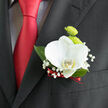 A refined boutonnière is pinned precisely on the lapel of a dark, tailored suit in this photograph, the flowers providing a luminous focal point against the deep fabric. The central bloom is a white orchid, its petals broad, smooth, and slightly cupped, as if gently holding the ambient light that might fall through a Brent Cross venue's windows before the ceremony begins. There is a whisper of creamy yellow at the heart of the orchid that adds depth, giving the flower a living, breathing presence. Behind it, round lime-green button chrysanthemums bring a cheerful burst of colour, their tight clusters of short petals forming tidy, textured spheres that contrast beautifully with the orchid's more fluid lines. Surrounding the main blooms, clusters of baby's breath scatter like tiny stars, each miniature white blossom perched on a fine stem, adding softness and a sense of airy movement. The arrangement is framed by deep green foliage, leaves polished and gently curved, grounding the design with a natural, garden-fresh feel reminiscent of local Brent Cross green spaces. At the base of the boutonnière, the florist has wrapped the stems in a narrow ribbon of deep red satin, the fabric's subtle sheen matching the rich red tie visible just below. Together, the colours-white for purity, green for new beginnings, and red for love-create an understated but emotionally resonant palette well-suited to weddings, civil ceremonies, and formal celebrations across Brent Cross and neighbouring areas. The boutonnière feels carefully considered, a small but significant detail that will stay with the wearer from the first photographs to the last dance of the evening.
