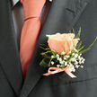 A refined, close-up study of a groom's boutonnière: a single peach-rose, small enough to be intimate yet full enough to command the lapel. The bloom's petals are softly unfurling, flushed with warm peach and blush tones that glow under soft daylight; each petal edge holds a subtle translucence, suggesting freshness and careful handling. Tiny clusters of white baby's breath (gypsophila) drift around the rose like miniature clouds, and slender, whispering green foliage creates gentle movement and contrast against the dark charcoal fabric. The boutonnière is bound neatly at the stem with a narrow, pale-pink satin ribbon, its slight sheen picking up tones from the matching peach tie worn over a light dress shirt patterned with a faint dark grid. The composition is photographed close enough to read texture - the rose's velvety surface, the crisp stitch of the jacket lapel, and the ribbon's satin smoothness - offering a tactile sense of the piece without needing to touch it. There's a soft, almost floral sweetness suggested by the image, a reminder of summer gardens and the quiet joy of a carefully chosen detail. Made in the spirit of local weddings and events around Brent Cross and nearby Golders Green, this boutonnière reflects the kind of small, considered craftsmanship we create daily at Brent Cross Flowers - a tiny emblem of ceremony, lovingly prepared to sit against a groom's heart.