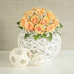 A hand-gathered dome of peaches and apricot roses rests atop an open, white-woven orb, its soft palette speaking of gentle celebration in the heart of Brent Cross. The roses are lush and abundant, each bloom shaped into a delicate spiral whose inner petals hold a whisper of cream while outer edges warm into apricot. Interspersed among them, clouds of white gypsophila drift like breath, their tiny blossoms catching light and lending an ethereal, almost veil-like effect. Sprays of deep green foliage provide contrast and structure, their cool tones grounding the softness of the petals and introducing a subtle, leafy fragrance.

The spherical rattan vessel has a contemporary, airy architecture: the white painted lattice throws soft shadows and lifts the floral dome visually, so the composition reads as both sculpture and bouquet. Sunlight glancing across the arrangement gives a pale golden glow, enhancing the peach hues and the powdery texture of the gypsophila. Perfect for a wedding, anniversary, or calm home styling, this piece conveys tenderness and refined craftsmanship - a design familiar to florists working around Hendon Way and the Brent Cross Shopping Centre. Prepared with careful hands and attention to seasonal freshness, this arrangement is available for local delivery by Brent Cross Flowers and makes a quietly confident statement of care and new beginnings.