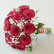 A classic romantic bouquet of red roses and white baby's breath fills the scene, as though poised on a sideboard in a Brent Cross apartment, catching the soft glow of afternoon light from a nearby window. The roses sit at the centre of the composition, each head full and rounded, their velvety crimson petals spiralling outward in intricate layers that suggest both opulence and tenderness. Nestled between and around them, airy clusters of gypsophila create a fine lacework of minuscule white blossoms, offering an ethereal contrast that brings a sense of delicacy and light to the deep reds. Touches of fresh green foliage peek out from beneath the blooms, framing the bouquet and adding a suggestion of movement, as if the stems have just been turned in the florist's hands in a studio close to Brent Cross Shopping Centre. The stems are gathered tightly into a neat, symmetrical shape, bound in a smooth ribbon that has been embellished with a line of dainty pearls, lending the handle a refined, almost heirloom quality reminiscent of bridal accessories chosen on nearby Finchley Road. The overall mood is one of quiet celebration and intimate devotion, like the stillness just before an engagement announcement at a restaurant around Hendon or an anniversary dinner at home. Imagining the sensory details, the roses likely release a softly sweet, familiar fragrance, grounded and comforting, while the gypsophila contributes a clean, almost airy freshness. Together they create a bouquet that looks and feels like a carefully composed love letter, ready to be delivered across Brent Cross and the surrounding neighbourhoods.