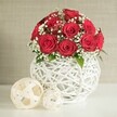 An elegant table arrangement of red roses fills this image, composed with the quiet sophistication you might find at a wedding breakfast in a Brent Cross hotel or intimate restaurant. At the centre, a lush, spherical bouquet of around a dozen to fifteen roses forms a dense, rounded mound. The blooms are a classic deep crimson, each flower open and layered with velvety petals that spiral tightly towards the centre, giving the arrangement a rich, almost plush appearance. Between the roses, delicate trails of white baby's breath thread their way through, their clusters of tiny blossoms providing a soft, cloud-like contrast that brightens the bouquet. Subtle flashes of green foliage peek out here and there, suggesting fresh, healthy stems beneath the flowers. The arrangement sits in an eye-catching white vase with a bulbous, globe-like shape. The vase is crafted from a network of slim, white strands that interlace to form an open lattice, reminiscent of finely woven wicker reimagined as modern sculpture. Light filters through its gaps, adding depth and shadow. On the smooth, white tabletop just in front and to the side of the vase, two cream-coloured decorative spheres rest, created from loosely woven, flat strips that echo the vase's texture. One sphere is noticeably larger and placed slightly forward, with the smaller ball set just behind, creating a pleasing visual balance. The backdrop is a softly blurred wall in a light beige or off-white, with a gentle, fabric-like texture that stays unobtrusive and calm. Together, the rich red roses, airy baby's breath, and woven white accents create a feeling of intimate celebration, making this an ideal representation of a wedding table arrangement crafted by a skilled florist serving Brent Cross and nearby North London areas.