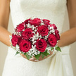 The image captures a close-up of a bridal bouquet held just in front of an ivory gown, the flowers arranged in a perfectly rounded dome of red and white. At the centre of attention are the red roses, their petals rich and velvety, unfolding in full, sculpted whorls that create a lush, almost plush surface across the top of the bouquet. Interlaced among the roses is a fine halo of baby's breath, each small white flower forming part of a soft, frothy ring that lightens the overall look and adds a dreamy, romantic texture. Just beneath the blooms, you can see a subtle edging of fresh green leaves, offering structure and a gentle hint of contrast, like a quiet frame around a vibrant painting. The bouquet appears expertly hand-tied, the stems carefully gathered as if ready to accompany a bride from a home near Brent Cross Station to a ceremony space in Hendon or Golders Green. The interplay of crimson, white, and green feels balanced and serene, evoking tradition without feeling old-fashioned. It is easy to imagine the fragrance: the familiar sweetness of red roses mingling with the clean scent of cut greenery as the bride moves through light-filled rooms. From the vantage of a local Brent Cross florist who knows the preferences of neighbourhood couples, this bouquet looks like a favourite choice for those wanting a bold yet classic statement, something that will photograph beautifully against a white dress and remain just as striking in memories as it is in the moment.