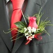 The image presents a refined wedding buttonhole pinned to a dark charcoal grey suit lapel, capturing the quiet poise of a groom preparing for vows somewhere near Brent Cross. At its heart lies a single cerise-pink rosebud, a deep, romantic shade of fuchsia with petals that curl inward in a tight spiral. The surface of the petals appears velvety and smooth, with soft highlights where the light touches their edges, suggesting freshness and care. Surrounding this central rose is a ring of white baby's breath; the tiny gypsophila flowers resemble a halo of miniature stars, each bloom adding a feather-light softness that contrasts beautifully with the darker tones of the suit. A thoughtfully arranged mix of greenery wraps the composition, pairing broad, shiny leaves with finer, feathery sprigs that give the boutonniere a sense of gentle movement, much like leaves swaying in the breeze near local Brent Cross parks and residential streets. At the base, a neatly tied bright red satin bow adds a playful yet elegant finishing touch, echoing the bold colour of the red silk tie that lies beneath the lapel. The tie falls smoothly over a subtly patterned grey dress shirt, whose quiet texture harmonises with the fine wool weave of the suit. The lighting is soft but clear, allowing every texture to stand out-the woollen lapel, the satin ribbon, the silky petals, and the airy baby's breath. The overall effect is one of understated romance and polished style, a boutonniere suited to grooms, groomsmen, or honoured guests attending weddings across the Brent Cross area, where small floral details speak volumes about love and intention.