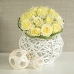 In this serene scene, a dome of soft yellow roses rises from a sculptural white vase, creating a centerpiece that feels tailored for a wedding or special dinner near Brent Cross. The roses are tightly packed, their velvety petals unfolding in layered spirals that give the bouquet a rich, cushion-like texture. Nestled among them are delicate stems of white baby's breath, their tiny blossoms flickering between the larger blooms like points of light, bringing an airy contrast and a touch of bridal charm. At the base of the flowers, a neat collar of smooth green foliage is wrapped with care, drawing a crisp line between the floral mass and the container below. The vase is distinctive: constructed from interlaced white strands that weave in and out to form an open, rounded silhouette with a gentle waist, it looks almost like a modern basket lantern, its gaps allowing light to filter through and dance across the tabletop. On the bright white surface nearby rest two spherical decorative accents in pale, natural tones, made from slender strips of wood or rattan twisted into open geometric shapes that echo the woven character of the vase. The background fades away into a gentle haze of beige and off-white, suggesting the soft walls of a contemporary home or event space in the Brent Cross neighbourhood, ensuring the viewer's attention remains on the flowers and their artful presentation. The arrangement exudes quiet sophistication and optimism, ideal for dressing tables at an intimate wedding, marking an anniversary meal, or adding a note of refined warmth to a local celebration arranged by Brent Cross Flowers.