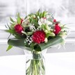 A luxurious red and white bouquet stands proudly in a clear cylindrical glass vase, its fresh green stems visible through the water like a tidy bundle of ribbons. At the heart of the arrangement, velvety deep red roses and a richly ruffled crimson carnation create a focal point of passion and warmth, their petals layered and gently unfurling as if just opened that morning. Around them, creamy white alstroemeria with soft speckles, dainty white chrysanthemums, and closed white lily buds bring a quieter grace, suggesting purity and the promise of blooms still to come. Wisps of gypsophila, or baby's breath, weave through the flowers like tiny stars, adding lightness and a cloud-like softness that balances the stronger reds. The bouquet is cradled by glossy dark green leaves, some artfully curled to form a neat collar at the base, while eucalyptus-style foliage and pointed leaves add texture and movement. The vase rests on a clean white surface, with a background that is softly blurred and bright, reminiscent of daylight streaming through a Brent Cross window on a calm morning. The gentle, even lighting highlights the sheen of each leaf and the depth of every petal, giving the bouquet a polished yet heartfelt character. This arrangement feels ideal for a romantic anniversary in a flat near Brent Cross Shopping Centre, a cheerful birthday surprise for an office on the North Circular, or a tender Mother's Day gesture delivered across the local area, carrying with it the quiet message of admiration, gratitude, and enduring affection.