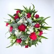 A richly detailed, top-down view of a circular hand-tied bouquet shows a dense, carefully layered mix of red and white flowers resting against a pure white background, as if just finished on the workbench of a Brent Cross florist. Deep crimson roses form the visual heart of the arrangement, some still in tight, velvety buds and others half-open, their spiralling petals hinting at romance and quiet luxury. Around them cluster ruffled fuchsia and ruby red carnations, their plush, feathery petals creating a sense of movement and adding a celebratory, almost party-like energy. Threaded between the reds are small, daisy-like white spray chrysanthemums, their clean, bright faces catching the light and lifting the palette, while shining white lily buds and blooms introduce a touch of serene elegance. Fine clouds of white gypsophila, or baby's breath, float in the spaces between stems, giving the bouquet a soft, misty halo that contrasts with the solid forms of the larger flowers. The whole design is cradled in an abundance of greenery: pointed dark green leaves from lilies or alstroemeria radiate outward to give structure, while rounded, silvery eucalyptus leaves weave through with a cool blue-green tone that feels modern and stylish, much like the contemporary homes around Brent Cross and nearby Golders Green. The lighting is bright and even, accentuating every texture-from the velvety rose petals to the waxy eucalyptus leaves-and making the bouquet feel freshly arranged and ready for delivery. The composition conveys joyful affection and heartfelt appreciation, perfectly suited to a Valentine's surprise, an anniversary dinner near the shopping centre, or a thoughtful birthday gift for someone living along Hendon Way, bringing a burst of colour, scent, and warmth into their day.