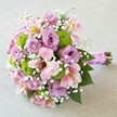 Vivid and romantic, this rounded bouquet-carefully arranged by Brent Cross's local florists-rests against a soft beige background, its colours glowing like a celebration beneath the glass canopy of the local shopping centre. The design features plump lavender roses at the centre, surrounded by playful lime green button mums that add a dash of contemporary style. Pink Alstroemerias, softly flared, introduce graceful movement with their butter-yellow and burgundy-speckled petals, while feathery clouds of gypsophila (baby's breath) create an ethereal effect throughout. Emerald green leaves peek from within, enhancing the bouquet's fullness and natural charm. Stems are tightly bound and wrapped in a plush fuchsia ribbon, adorned with pearl accents-a subtle, romantic detail. Ideal for Brent Cross weddings, spring gatherings, or heartfelt gifts delivered across the local area, this bouquet exemplifies the blend of elegance and freshness beloved by the NW4 community.