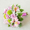 Resting on a neutral, slightly textured fabric that suggests linen or fine burlap, this bouquet is photographed from above at a gentle angle, allowing each flower to be seen clearly. Central to the design are classic soft pink roses, their petals layered and slightly cupped, spiralling inward with a serene, romantic beauty. Between the roses, lime-green button chrysanthemums form compact spheres, their tiny petals packed tightly together, offering a crisp contrast in both texture and colour that instantly brightens the arrangement. Pink alstroemeria add a sense of movement, their elongated petals flaring outward and decorated with intricate streaks of darker pink and hints of yellow, creating a hand-painted effect. Small clusters of baby's breath weave through the bouquet, their tiny white blossoms like scattered stars, adding airiness and softening the lines between each larger bloom. Around the outside, lush greenery in varied shades of green frames the central flowers, with leaves that appear smooth, slightly glossy, and gently curved, giving the design a fresh, garden-inspired feel. The stems are gathered together at the base and wrapped in a smooth magenta satin ribbon, which lies neatly and securely, its rich colour playing beautifully against the pink roses and green chrysanthemums. The light is soft and even, emphasizing the natural colours without harsh shadows. This bouquet would be perfectly at home as a bridal arrangement for a wedding near Brent Cross, a romantic gesture delivered to a nearby apartment overlooking the North Circular, or a thoughtful gift from Brent Cross Flowers to mark birthdays, engagements, and anniversaries in the local community.