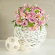 Captured in a close-up that highlights every texture, this wedding-perfect floral centerpiece feels like it belongs on a beautifully dressed table in a Brent Cross townhouse. The bouquet forms a gentle, domed shape, densely packed with roses in delicate lavender and rich pink, their petals curling outward in layered spirals that suggest softness at a glance. Nestled among them are pale pink alstroemeria, or Peruvian lilies, their trumpet-shaped blooms brushed with fine freckles and streaks of deeper pink and apricot. Threaded through the pastel tones are vivid lime green button chrysanthemums, compact and rounded, adding a lively pop of colour and a contemporary twist. Wisps of white baby's breath run between the main flowers like fine lace, softening transitions and giving the arrangement a romantic, airy quality. You can just see hints of dark green foliage at the base, supportive but discreet, framing the flowers rather than dominating them. The entire dome of blooms sits in a spherical white vase crafted from woven wicker, its open lattice structure creating a beautiful contrast of light and shadow and giving the design an almost sculptural presence. On the same smooth white surface, two smaller woven balls in a pale, natural tone rest casually to one side, echoing the texture and adding a touch of organic décor. The neutral, off-white background keeps the focus on the flowers, reminiscent of a softly painted wall in a Brent Cross flat or local wedding venue. The mood is romantic but clean-lined-perfect for a registry ceremony table, a top table display, or an elegant gift delivered locally for a quiet celebration of love.