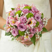 This elegant bridal bouquet blends shades of lilac, pink, crisp green, and white into a design that feels both contemporary and timeless, much like a quiet corner of Brent Cross just away from the busy roads. At the heart of the arrangement, dusky mauve roses bloom in layered spirals, each flower lush and full, with velvety petals that seem to hold the light. Around them, stems of gypsophila scatter tiny, star-like blossoms, forming a delicate haze that softens the outline of the bouquet, adding a whispery, almost ethereal texture. Bright chartreuse button chrysanthemums peek through the roses and baby's breath, their neat, rounded forms and closely set petals bringing a playful, tactile element that invites the fingers to explore. Pink alstroemeria weave through the design, their elongated petals gently flaring outward, decorated with fine streaks and speckles that add grace and subtle rhythm to the composition. Surrounding the blooms, a collar of rich greenery frames the bouquet, the leaves smooth and glossy, some gently arching, others providing structure and depth, giving the impression of fullness without weight. The lighting in the image is soft and natural, enhancing the pastel tones of the roses and the brightness of the greens, as if the bouquet were photographed by a sunlit window near Hendon Way or close to the Brent Cross Shopping Centre. There is an impression of fresh fragrance-roses carrying a gentle perfume, mingling with the clean, herbal note of chrysanthemum foliage. Suitable for a wedding ceremony at a local synagogue, church, or hotel venue, this bouquet captures the essence of partnership and celebration, each flower carefully positioned to create a sense of harmony, balance, and heartfelt joy.