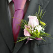 This carefully composed boutonnière appears against a dark, tailored suit, its soft colours and precise structure creating a graceful contrast ideal for a Brent Cross wedding or prom. At its centre is a pale pink rose, just opening, each petal smooth and velvety with a gentle blush that deepens toward the core. The rose feels both delicate and confident, like a quiet smile before walking into the ceremony. Around it, well-trimmed greenery forms a clean, polished collar, the leaves lying close and flat so the arrangement sits comfortably on the lapel. Among these, clusters of tiny white gypsophila blooms scatter delicately, giving the impression of a light, floral mist that softens the outline and adds a hint of whimsy. Tucked near the lower portion of the design, a green button chrysanthemum introduces a vivid pop of lime, its tight, rounded petals arranged in a perfect dome that hints at modern floral styling. A slender blade of foliage extends beside the main flowers, directing the eye downwards to the stems, which are bound together in a soft mauve ribbon. The ribbon is tied with care, its ends trailing just enough to suggest movement, like the swish of a gown on the way into a reception near Brent Cross station. The mauve tone offers a subtle, sophisticated link between the green and pink hues, holding the palette in harmony. You can almost imagine the faint scent of the rose and the cool touch of the satin ribbon as an experienced florist from Brent Cross Flowers gently pins the piece in place, ensuring it rests securely close to the heartbeat of the wearer on this memorable day.