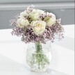 A close-up photograph shows a serene white and lavender bouquet glowing softly against a pale, blurred background, as if placed on a bright windowsill in a quiet Brent Cross home. At the heart of the design are several creamy white roses, their petals layered and gently unfurling, with the faintest suggestion of pale buttery yellow deep in the centres that hints at absolute freshness. Each rose head is full and velvety, the kind of bloom that a skilled florist selects carefully at just the right stage so that it will open beautifully over a few days. Around them, like a mist, spreads an abundant halo of tiny lavender and lilac-hued statice flowers. Their papery, cloud-like clusters create a soft, romantic haze, adding both fullness and a delicate, almost featherweight texture that contrasts with the smooth, voluptuous rose petals.
The bouquet is gathered into a clear glass vase with a distinctive crackled or ice-like texture, the surface catching the light in small, sparkling lines that feel almost frosted. Through the water inside, the straight green stems are clearly visible, cleanly cut and neatly arranged, suggesting professional preparation and very recent conditioning. Everything rests on a smooth, white tabletop that reflects a little of the daylight and throws a gentle shadow beneath the vase, reinforcing the impression of calm and balance.
The overall mood is peaceful and understated, like a quiet morning walk through the greenery near Brent Cross or along local residential streets before the city fully wakes. This arrangement feels ideal for a thoughtful sympathy gesture, a gentle get-well wish, or to bring a sense of calm elegance to a bedroom or study, its soft colours and relaxed shape offering comfort without ever feeling overpowering.