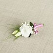 A delicately crafted wedding boutonniere rests at a gentle diagonal on a softly textured, light beige fabric that looks like finely woven linen, its natural oatmeal tones warming the whole scene. At the heart of the design sits a fully opened white lisianthus bloom, its ruffled petals layered like soft silk, forming a rounded cup of creamy white that feels both romantic and serene. Just below and to the left, two slender eustoma buds in pale cream and green nestle close, suggesting blossoms that will open later in the day, much like the unfolding of a new marriage. Tiny tufts of white gypsophila, or baby's breath, are interwoven between the main flower and buds, their cloud-like clusters adding an airy, almost ethereal lightness. Slender, grass-like blades of fresh green foliage sweep gracefully towards the lower left, creating a sense of movement, while a single, broader dark green leaf anchors the composition behind the main bloom, giving the arrangement depth and structure. The stems are neatly gathered and bound with a vivid fuchsia satin ribbon tied into a neat, jaunty bow, its bright pink sheen catching the light and providing a joyful contrast to the calm whites and greens. The soft, even lighting highlights every petal edge and thread of fabric, lending the image a peaceful, intimate mood, as if captured just before the ceremony begins. It feels perfectly suited for a groom's lapel at a Brent Cross registry office wedding, a prom on a quiet residential street off the North Circular, or any special occasion where a simple yet refined floral detail can carry the quiet excitement of the day.