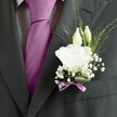 A sharply focused close-up of a groom's charcoal grey suit jacket, captured just at chest level so the viewer sees only the refined fabric, the knot of his tie, and the detailed boutonnière pinned to his left lapel. The suit, likely a fine wool with a subtle weave, sits smoothly over a pale grey and white patterned dress shirt, each line pressed and immaculate, as you might expect at a smart Brent Cross wedding near the shopping centre or Hendon Way. Between the lapels, a rich magenta or deep fuchsia silk tie glows against the cooler tones, perfectly knotted with a sleek, light-catching sheen. Nestled against this formal backdrop is a carefully crafted buttonhole: a single, fully open white lisianthus or rose-like bloom with soft, ruffled petals forming a layered cup, accompanied by two slender ivory buds leaning protectively above it. Around them, clouds of tiny white gypsophila blossoms create a halo of texture, while fine, airy greenery arcs lightly outward, adding gentle movement and freshness. At the base, a single broad green leaf curves neatly around the stems, which are bound with a narrow mauve or pinkish-lavender ribbon tied into a small, precise bow that echoes the colour of the tie. The overall mood is calm, romantic elegance, ideal for describing groom or groomsmen attire chosen for a ceremony in one of the nearby North London venues, where every detail is considered and the floral work feels both personal and expertly composed.