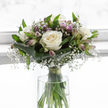 A thoughtfully crafted sympathy bouquet sits in a clear glass vase, the flowers bathed in pale, diffused light that lends them a gentle radiance, as if they were placed by a window in a quiet Brent Cross home on a still afternoon. At the centre of the composition, large white roses are fully open, their velvety petals layered in generous spirals that communicate innocence, memory, and quiet grace. Surrounding these roses, fine sprays of gypsophila create a soft halo, each tiny, pristine blossom contributing to a cloud-like effect that softens the bouquet's contours and lends an almost whispered presence. Interspersed within this airy surround, elegant stems of alstroemeria rise with elongated buds and partially open blooms, brushed in delicate tones of lavender, pale purple, and muted lilac, some petals lightly striped in a way that adds gentle character without disturbing the arrangement's calm mood. The base is embraced by broad, dark-green leaves, meticulously folded and wrapped to form a structured, almost protective collar around the blooms, grounding the design and emphasizing its rounded form. Finer green leaves extend upward, adding a sense of life and natural movement. Inside the cylindrical vase, the long stems are clearly visible, fresh and upright in clear water, secured neatly with a slim green binding that keeps them perfectly aligned. The overall feel is one of serene composure and heartfelt sincerity, the kind of arrangement Brent Cross Flowers might create for a condolence visit, funeral, or remembrance gathering near the shopping centre or along Hendon Way, when a quiet, beautiful floral tribute is needed to honour someone dearly missed.
