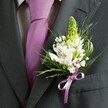 A close-up, high-angle photograph shows the upper torso of a man dressed for a formal occasion somewhere in Brent Cross, his charcoal grey suit jacket filling the frame. The fabric looks smooth and expertly tailored, with a sharp lapel and clean shoulder line that catch soft, natural light, perhaps by a window overlooking the shopping centre or nearby leafy streets. Peeking from beneath the open lapels is a crisp, pale dress shirt with a subtle grid pattern, its cool tones perfectly complementing a sleek lavender tie that falls straight and neat down the centre of his chest. Pinned carefully to the left lapel is the true focal point: an elegant wedding boutonniere, created with the care of a local florist. A tall, pale green spiky flower bud rises at the top, echoing the shape of hyacinth or Muscari, its unopened florets suggesting anticipation and new beginnings. Around it cluster white Alstroemeria blooms, each petal brushed with fine maroon streaks and a hint of gold at the stamens, like delicate strokes of paint. Tiny white clouds of baby's breath nestle between the flowers, their airy heads softening the overall shape. Slim, grass-like blades arc gracefully among broader, darker green leaves, adding movement and texture against the dark suit. At the base, a satin lavender ribbon is tied into a neat bow, perfectly matched to the tie, drawing the eye to the craftsmanship. The scene feels calm and expectant, as though captured just before a wedding ceremony in one of the venues around Brent Cross, the fresh florals quietly marking a milestone moment.