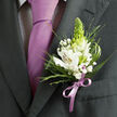 This image focuses tightly on the left lapel of a dark charcoal grey suit, revealing the kind of precise detail a local Brent Cross florist would perfect for a wedding party gathering near Hendon Way. The suit itself is crisply pressed, its lapel smooth and slightly matte, forming a strong frame for the floral work. Nestled against the fabric, a white alstroemeria boutonniere opens gracefully, each lily-like bloom showing delicate reddish-brown speckles and fine violet striations across creamy petals, with golden-yellow throats that seem to glow. Standing tall in the center, a fresh, lime-green tapered spike of buds adds height and movement, suggesting new beginnings and quiet excitement. Surrounding the main flowers are wisps of gypsophila, their miniature white blossoms forming a soft halo, along with airy, grasslike sprigs and more substantial deep green foliage that fans outward in natural arcs. The greenery ranges from slender, needle-like stems to broader leaves with a gentle sheen, creating layers of depth against the dark wool of the jacket. Beneath the flowers, a satin ribbon in pale lilac is wrapped and tied into a compact bow, its color perfectly mirroring the smooth lavender tie that hangs down over a finely patterned, almost gingham-like light shirt. The lighting is gentle and even, picking up the subtle shine of the tie and ribbon while leaving the suit slightly in shadow, making the boutonniere the clear focal point. The overall impression is of calm, refined celebration: a carefully coordinated buttonhole ready for a ceremony, formal dance, or special event in and around Brent Cross, prepared with the quiet confidence of an expert florist.