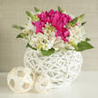 This image shows a beautifully composed floral basket, poised on a smooth white surface as if styled for a wedding table near Brent Cross. In the centre, an exuberant cluster of fuchsia blooms - plump and full, like peonies at their most lavish - captures the gaze. The petals overlap in soft, voluminous layers, shifting between bright magenta and deeper berry tones where the light falls away. Encircling this intense core, creamy white alstroemeria unfold in a gentle ring. Their slender petals, each adorned with fine speckles and streaks in soft pink and pale gold, create a halo of brightness that contrasts delicately with the vivid centre. Fresh green leaves form a crisp base, and several slim stems with unopened buds rise between the blossoms, suggesting new flowers on the verge of opening, much like the subtle seasonal changes you notice along the streets linking Brent Cross Shopping Centre to the surrounding residential avenues. 

The arrangement is housed in a gleaming white wicker basket, round and deep, its sides tightly woven in a modern lattice. The weave is both decorative and functional, giving the basket a sculptural quality that would sit comfortably on a stylish dining table or console. Beside it, two creamy, woven spheres rest quietly, their texture harmonising with the basket and lending a sense of curated design to the entire scene. Soft, even lighting washes across the flowers and wicker, highlighting the interplay of colour and form while keeping the mood calm and uncluttered. This bouquet feels ideal for wedding celebrations, engagement parties, or milestone anniversaries celebrated in and around Brent Cross, conveying polished elegance and heartfelt warmth. As you imagine the composition, you can almost catch the whisper of a fresh, green fragrance that enhances its serene yet joyful presence.