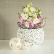 Bringing a breath of renewal to a Brent Cross home, this captivating still life presents a handcrafted white spherical vase-its open rattan or branch weave as light and intricate as the décor found in boutique flats along Prince Charles Drive. An abundant arrangement overflows from the vase: creamy lisianthus and soft yellow eustoma show off their multi-layered, ruffled petals, interspersed with vertical stems of pale pink matthiola that contribute both height and lushness. At the foundation, tiny purple filler flowers-perhaps asters or decorative alliums-create a lively undercurrent, bordered by a band of rich, luxuriant greenery. To the vase's left, two sculpted orbs fashioned from pale natural woven rattan add a tactile, organic accent, enhancing the handcrafted theme and bringing harmony to the overall composition. The bright white table reflects daylight that streams in softly, evoking the peaceful ambiance of a spring afternoon nearby Claremont Park. Behind, a subtly textured, warm beige wall gives the whole scene a neutral yet welcoming frame, allowing the gentle spectrum of pastel blooms to stand out with clarity and grace. This arrangement embodies the artistry and local flair that Brent Cross residents appreciate-ideal for wedding banquets, anniversary brunches, or simply welcoming the season's fresh palette into the heart of the home.