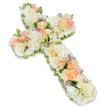 A serene and lovingly crafted floral cross is shown in its entirety, set against a bright white background that emphasizes its gentle colours and refined form. The cross shape, clearly recognisable as a Christian symbol, is softened by a complete covering of closely packed blossoms, giving it a pillowed, almost cloud-like appearance. The foundation consists primarily of white and creamy ivory flowers, with velvety roses and rounded hydrangea blooms creating a full, plush texture that suggests both comfort and reverence. Running through this pale base, clusters of roses in soft peach, apricot, and blush pink add warmth and emotional depth, their layered petals unfurling in graceful spirals. Smaller spray roses and delicate filler flowers fill in the spaces, ensuring no gaps interrupt the flow of petals. Scattered sparingly across the arrangement are tiny hints of lavender or lilac flowers, which introduce a whisper of purple that enriches the pastel scheme. At the edges, a fine border of fresh green foliage peeks out from beneath the blossoms, defining the outline of the cross while preserving its rounded, gentle silhouette. The lighting is even and clear, casting a light shadow beneath the arrangement and giving it a three-dimensional presence against the otherwise minimal background. This floral cross feels perfectly suited to a funeral, wake, or church service in Brent Cross or nearby areas along Hendon Way, created by local florists who understand the importance of a quiet, dignified tribute. It conveys sympathy, faith, and remembrance, offering a visual expression of comfort for families coming together to honour and say goodbye to someone dear.