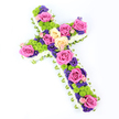Photographed on a clean white background, this floral cross is presented at a gentle diagonal angle, giving the sense of a carefully placed tribute ready for a service. Its outline is formed by a thick frame of white spray chrysanthemums, each small bloom nestled closely together to create a plush, cloud-like border that softens the strong, Christian cross shape. Just beneath this fringe of white runs a narrow band of light-green ribbon or folded foliage, its carefully pleated texture adding structure and a tender hint of verdant colour that subtly lifts the design. Within this border, the centre of the cross is filled with a vibrant combination of flowers. Large roses in vivid fuchsia pink and creamy pale yellow, some shading toward peach, are thoughtfully spaced along the arms and down the centre, their open, velvety petals conveying warmth and affection. Between them, rounded clusters of lime-green button chrysanthemums appear like tiny pom-poms, introducing lively texture and a fresh, modern note. Interwoven through the larger blooms are smaller, deep purple and violet blossoms-likely lisianthus or statice-that nestle into gaps, deepen the colour palette, and create a sense of quiet reflection. The lighting is bright and even, highlighting each element so the tribute feels crisp and newly arranged, as though prepared that morning in a Brent Cross florist's studio. The overall mood is one of respectful remembrance balanced with gentle optimism, making this floral cross a fitting choice for funerals, memorial services, or cemetery tributes in and around Brent Cross, where families may wish to honour loved ones with a symbol of faith, peace, and enduring love.