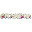 The photograph focuses on a graceful GRANDMA funeral tribute, laid out in a calm, horizontal line that would sit beautifully at a memorial service in Brent Cross or nearby Barnet. Each letter is crafted entirely from white chrysanthemums, arranged so densely that the surface appears like a smooth, floral cushion. Their petals are rounded and soft, overlapping to create gentle shadows and a feeling of depth within the pure white. A slim border of lilac ribbon traces the outer edge of every letter, its pastel tone adding a quiet touch of colour and refinement. The ribbon is carefully secured, following each curve and angle with precision, giving the tribute a polished, finished look that reflects the skill of the florist. At three points-the first G, an A in the centre, and the final A-small floral sprays add warmth and personality. These mini bouquets contain bright pink gerbera daisies, delicate pink sprays of roses, and sprigs of glossy green foliage, which spill gracefully over parts of the lettering. The pink tones bring to mind affection, laughter, and treasured memories, while the greenery introduces a natural freshness reminiscent of local green spaces around Brent Cross. Gentle, even lighting ensures that the individual blooms, leaves, and the sheen of the ribbon are all clearly visible, enhancing the sense of purity and calm. The tribute as a whole feels affectionate and sincere, a loving floral expression meant to honour a grandmother's life, presence, and the enduring bonds she leaves behind.